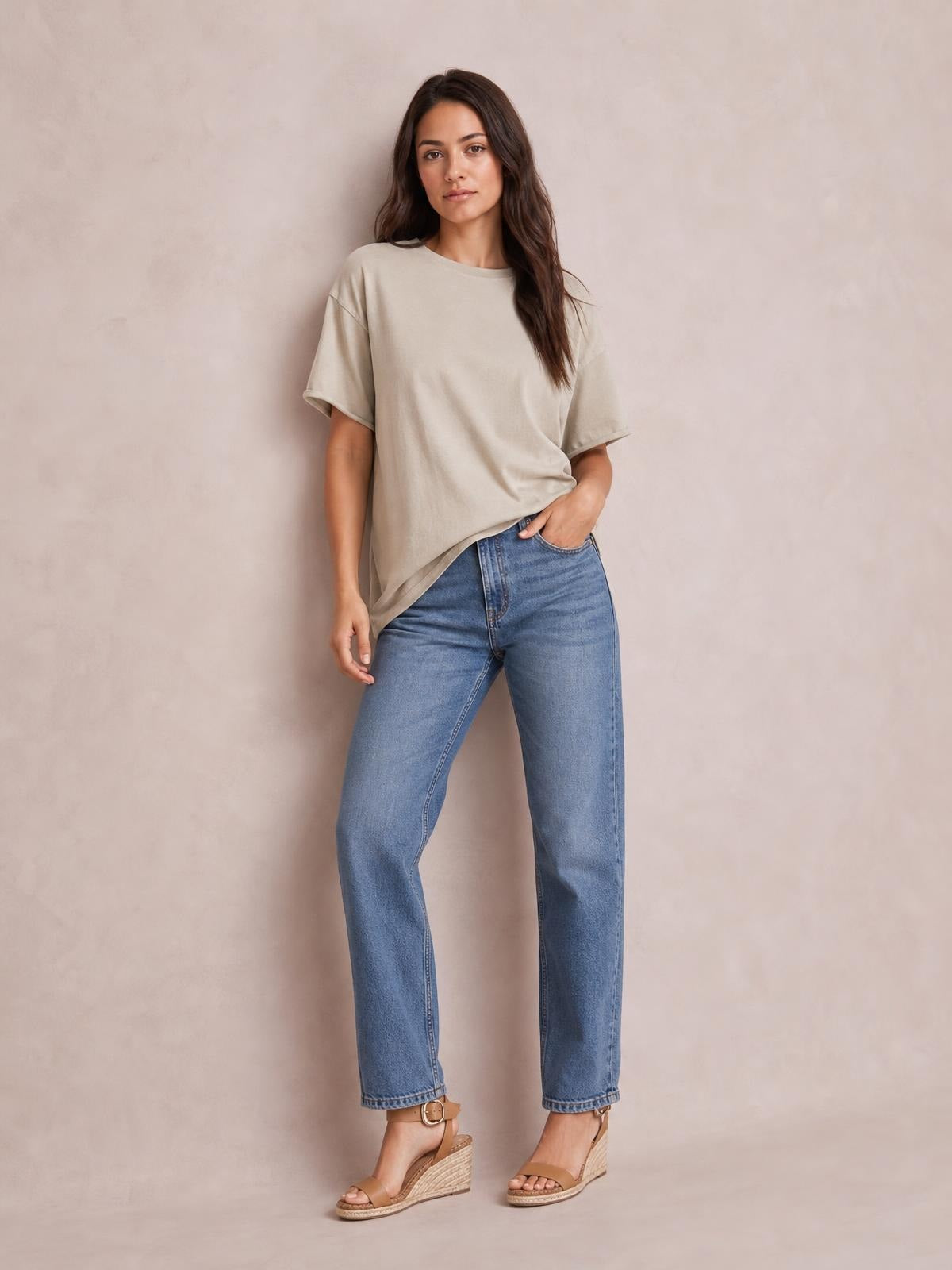 Woman wearing a beige exposed seam round neck half sleeve T-shirt with a relaxed silhouette, standing casually with one hand in her jeans pocket.