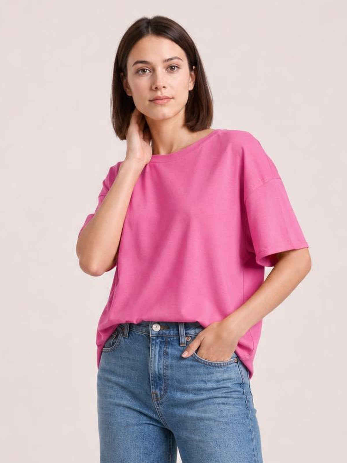 Model wearing a bright pink exposed seam round neck half sleeve T-shirt with a relaxed silhouette, posing with one hand at her neck and the other in her jeans pocket.