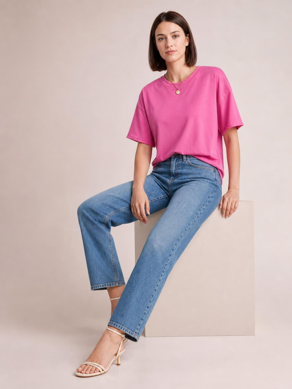 A seated model wears a pink exposed-seam round-neck half-sleeve T-shirt with a relaxed, slightly oversized silhouette and dropped shoulders.