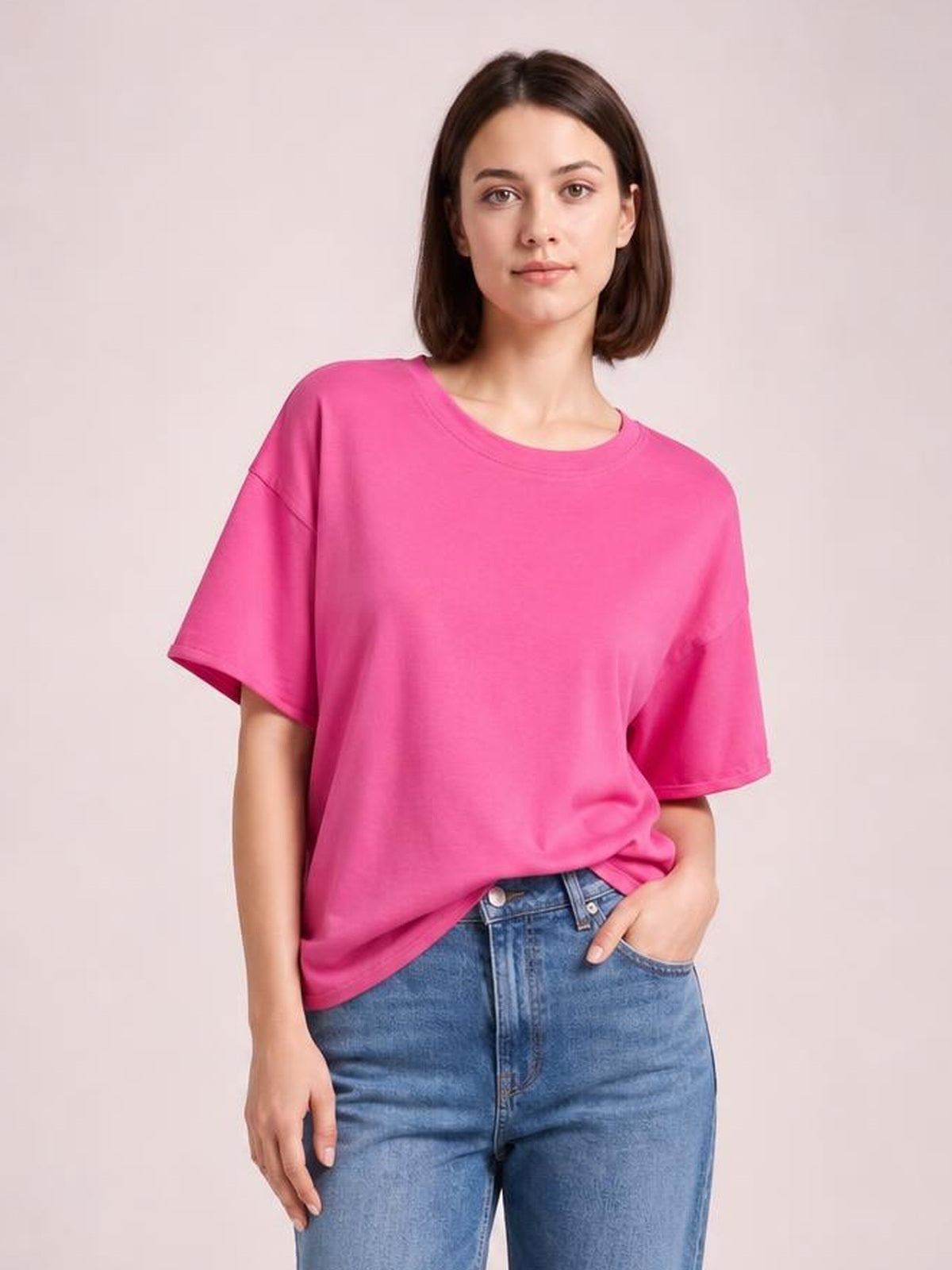 Model wearing a bright pink exposed seam round neck half sleeve T-shirt with a relaxed silhouette, standing with one hand in her jeans pocket.