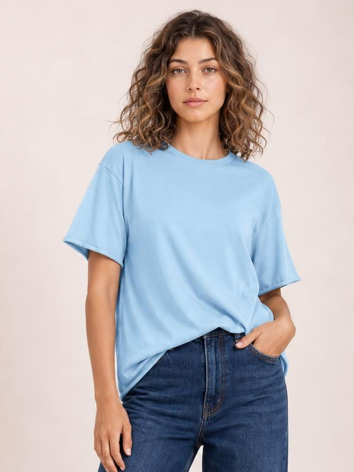 A model wears a light blue exposed seam round neck half sleeve T-shirt with a relaxed silhouette, standing front-facing with one hand in her jeans pocket.