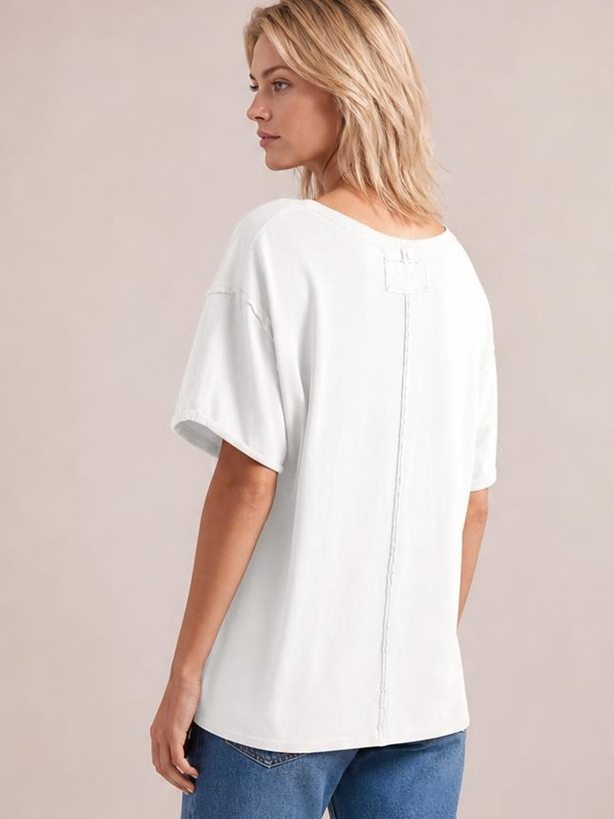 A model turns to show a white exposed seam round neck T-shirt with relaxed half sleeves, an oversized silhouette, and a visible center-back seam.