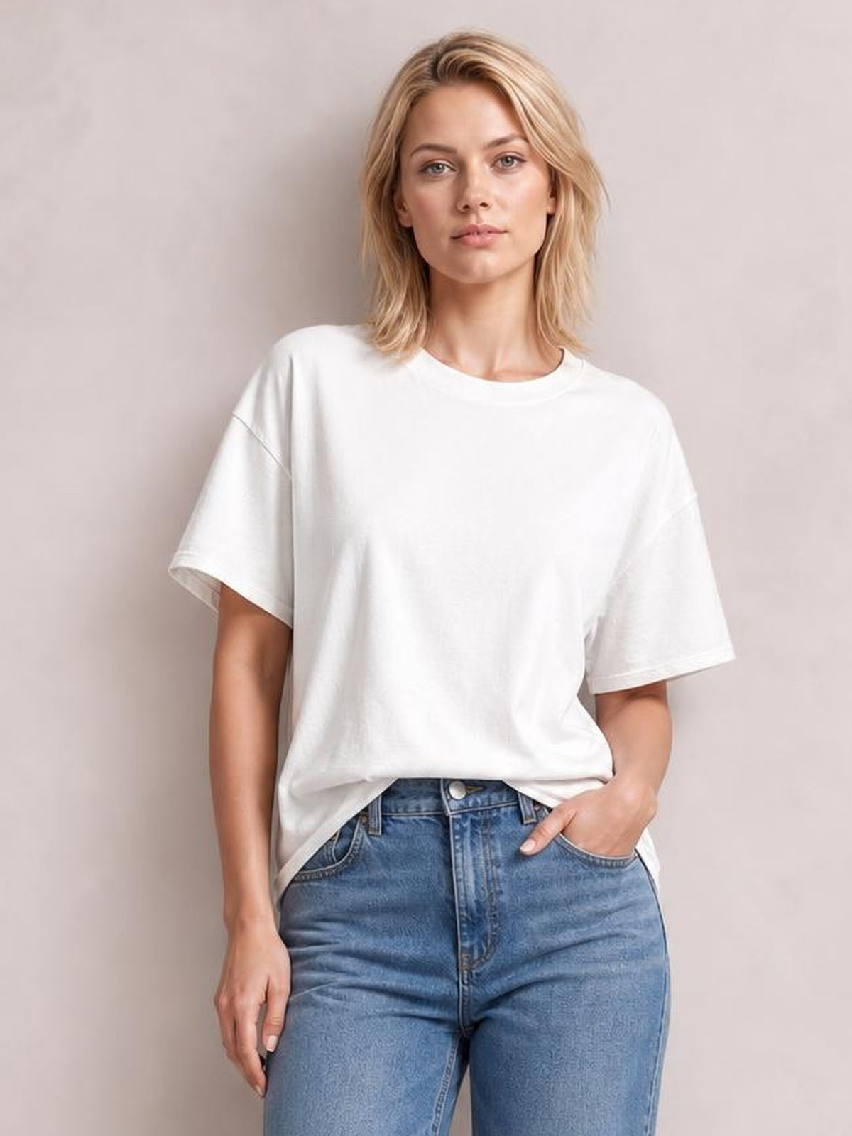 Model poses in a white exposed seam round neck half sleeve T-shirt with a relaxed oversized silhouette, slightly dropped shoulders, and a front tuck into blue jeans.