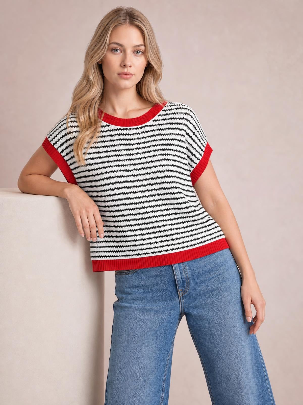 Model leans with one hand on a ledge wearing a white and black striped sweater with red ribbed round neck, cap sleeves, hem, and relaxed silhouette.