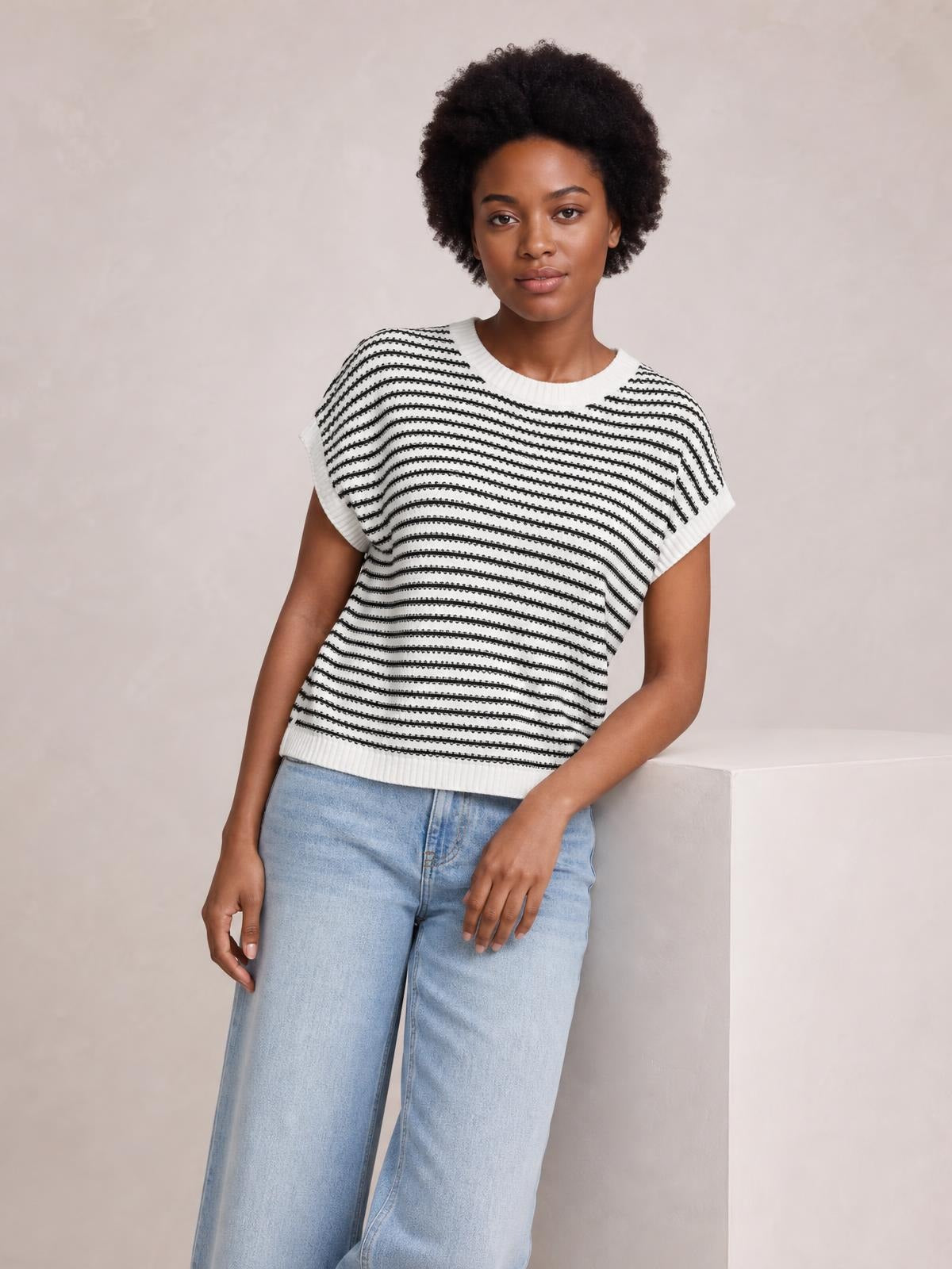Model wearing a white striped round neck cap sleeve sweater with ribbed trim and a relaxed silhouette, leaning casually with one hand resting on a ledge.