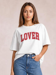 A model wears a white Lover round neck half sleeve T-shirt with a relaxed silhouette, red varsity-style lettering, and one hand in her jeans pocket.