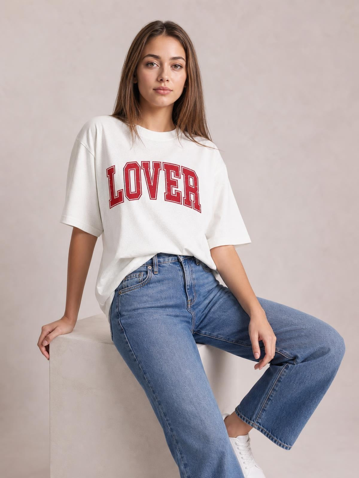 A model sits wearing an oversized white round neck half sleeve T-shirt with bold red “LOVER” lettering, a relaxed silhouette, and a casual draped hem.