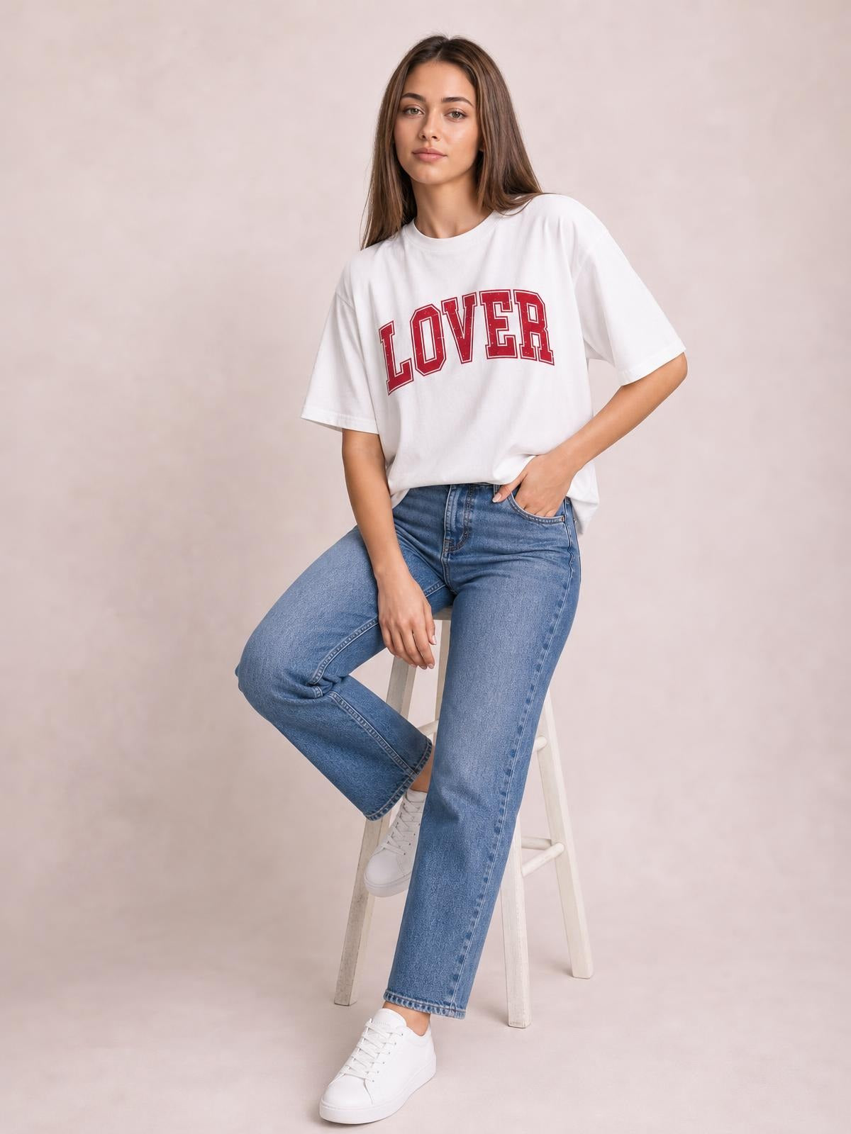 A model sits on a stool wearing a white Lover round neck half sleeve T-shirt with a relaxed silhouette and bold red front lettering.
