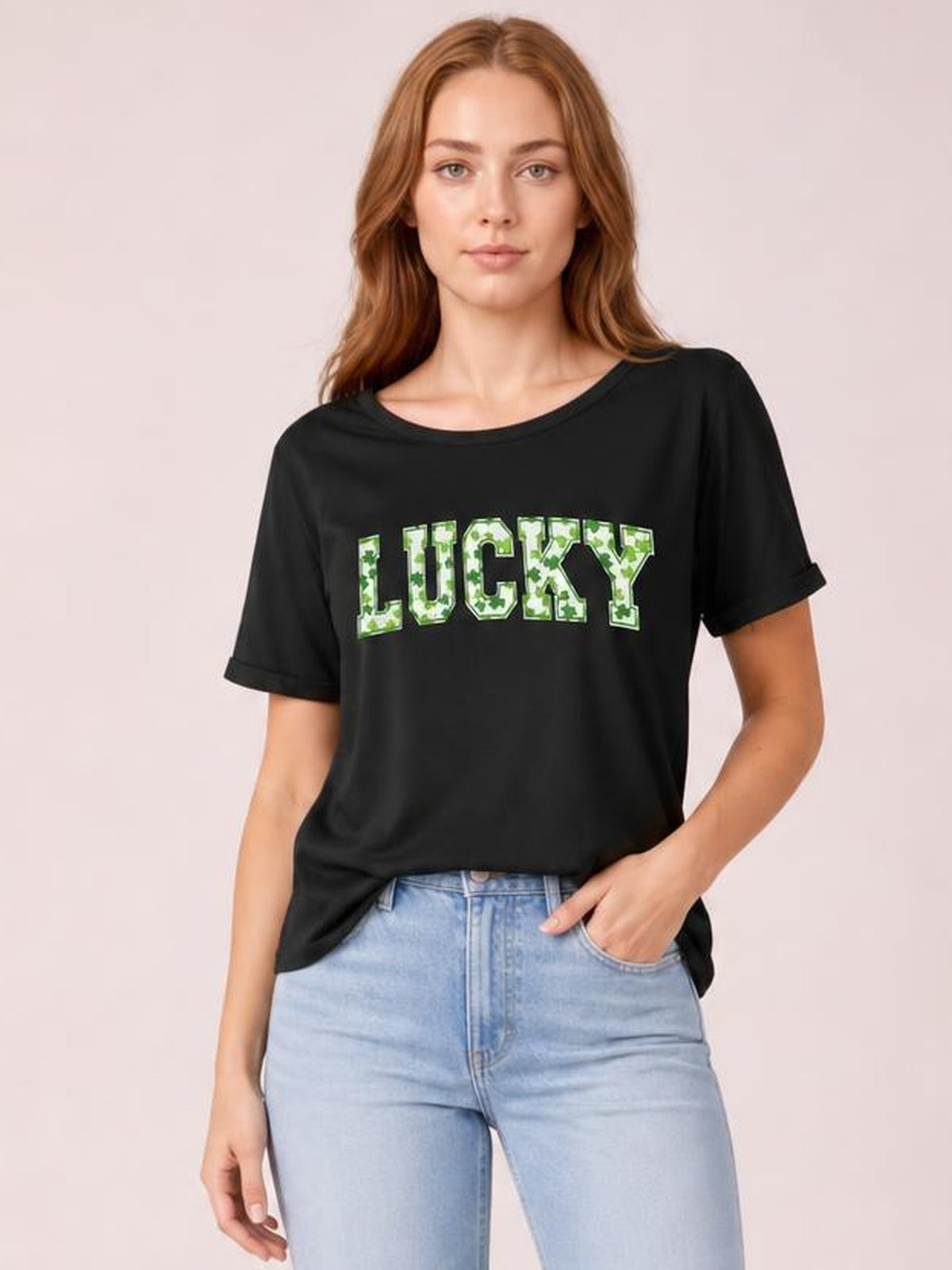 Model poses with one hand in her jeans pocket, wearing a black short sleeve T-shirt with rolled cuffs, a crew neckline, and bold clover-print “LUCKY” lettering.