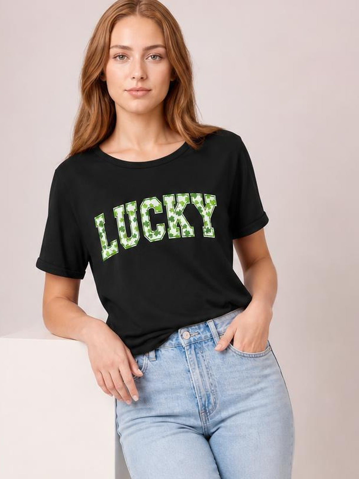 Model wearing a black short-sleeve LUCKY T-shirt with green clover-letter graphics, crew neckline, and relaxed silhouette, posing with one hand in her jeans pocket.