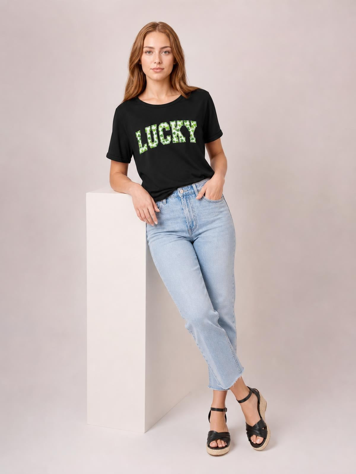 A model leans against a pedestal wearing a black short-sleeve T-shirt with bold green-and-white LUCKY clover lettering and a relaxed silhouette.