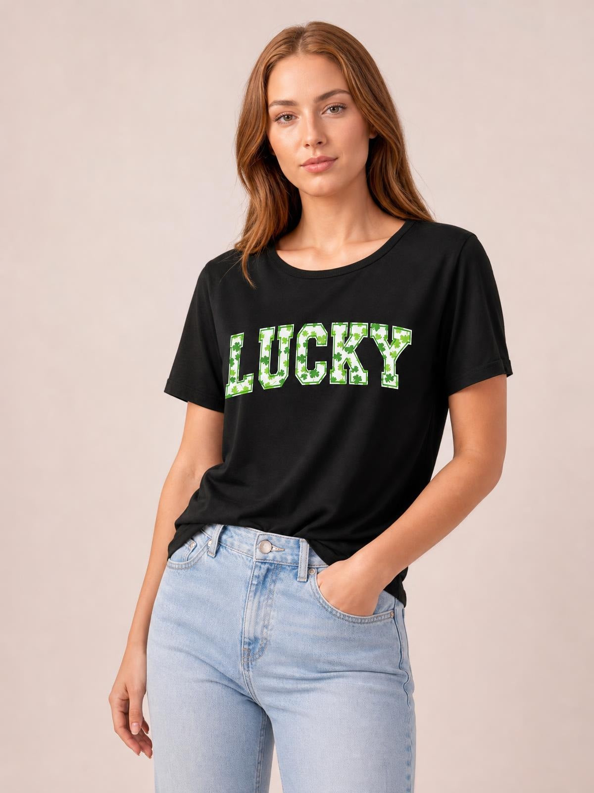 Model wearing a black short-sleeve T-shirt with a relaxed silhouette, crew neckline, and bold green-and-white “LUCKY” clover lettering, posing with one hand in her jeans pocket.