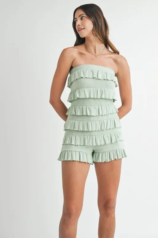 Model poses with hands behind her back in a pale green pointelle knit tube romper with a fitted silhouette and multiple horizontal ruffled tiers and shorts.