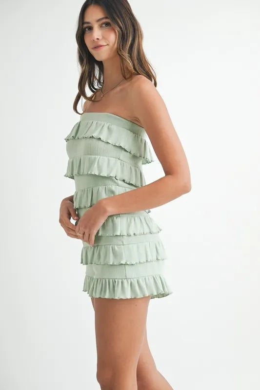 Model stands in a side pose wearing a mint green pointelle knit tube romper with a strapless neckline, fitted silhouette, and layered ruffled construction.