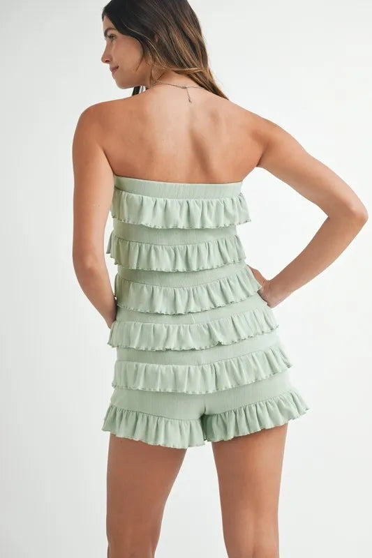 Model poses in a pale green pointelle knit tube romper with a strapless neckline, tiered ruffle accents, and a playful shorts silhouette.