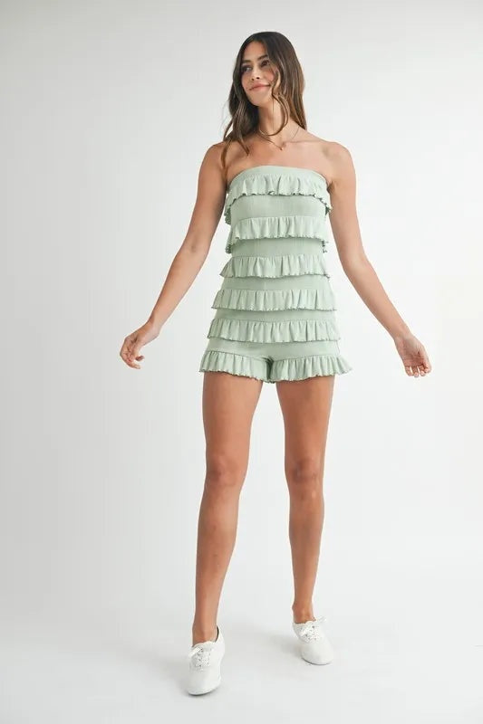 Model wears a pale sage pointelle knit tube romper with tiered ruffled rows, a strapless neckline, and a relaxed short silhouette while standing with arms out.