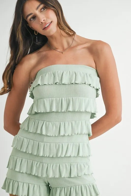 Model wearing a sage green pointelle knit tube romper with a fitted silhouette, layered ruffled tiers, and hands posed behind her back.