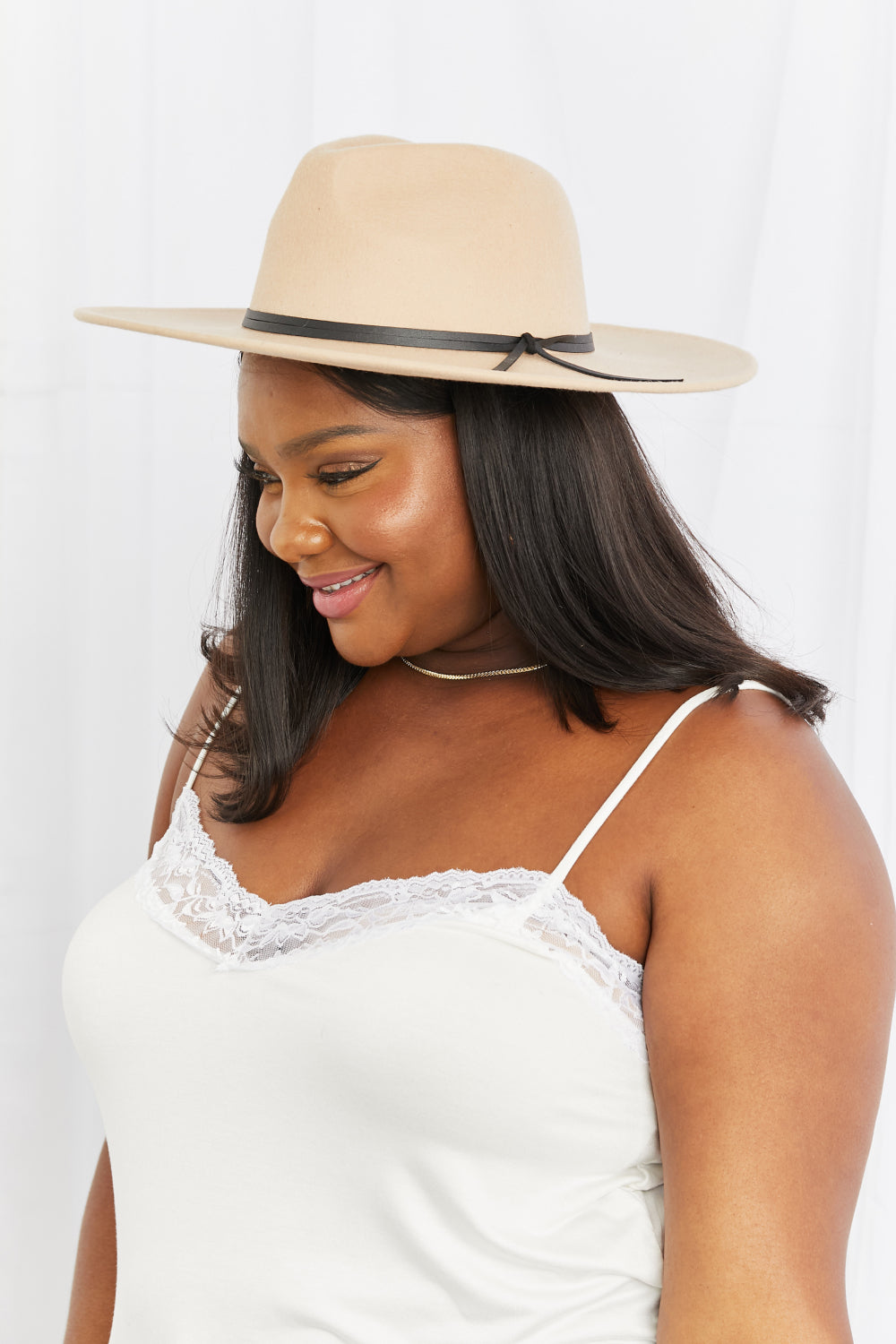 A smiling model looks down while wearing a beige fedora with a softly structured crown, wide brim, and contrasting black band with tied detail.