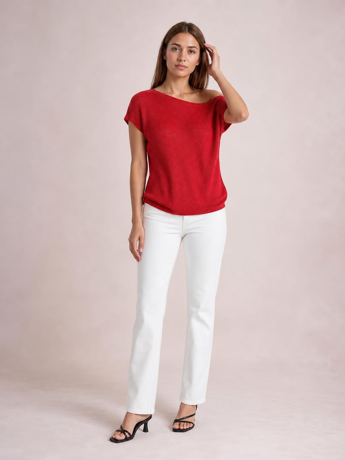 Model wearing a red boat neck short sleeve knit top with a relaxed silhouette, standing in white pants with one hand touching her hair.