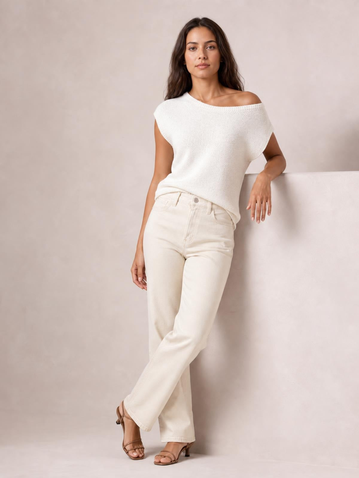 Model wears a white boat neck short sleeve knit top with a relaxed silhouette and wide neckline, leaning casually with one leg crossed.