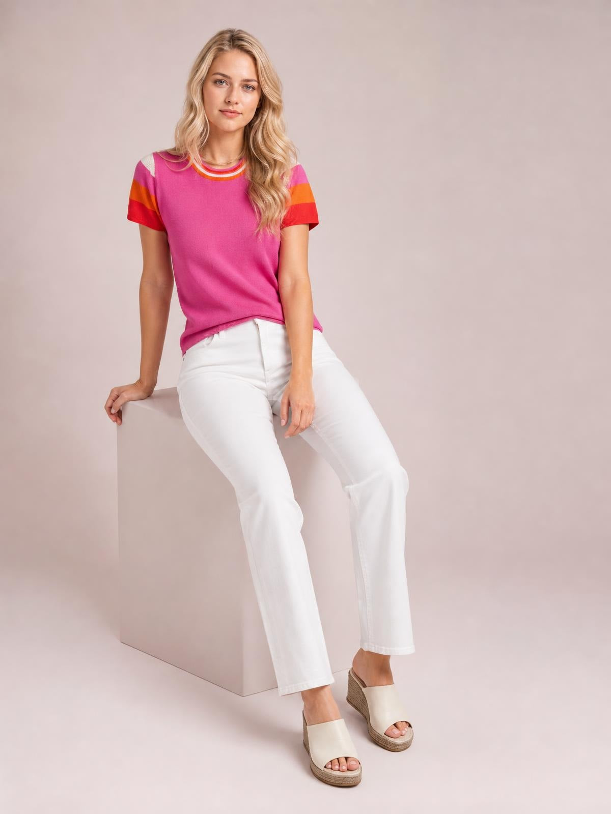 Model seated and wearing a bright pink contrast knit top with a round neck, short striped sleeves, and a relaxed straight silhouette.