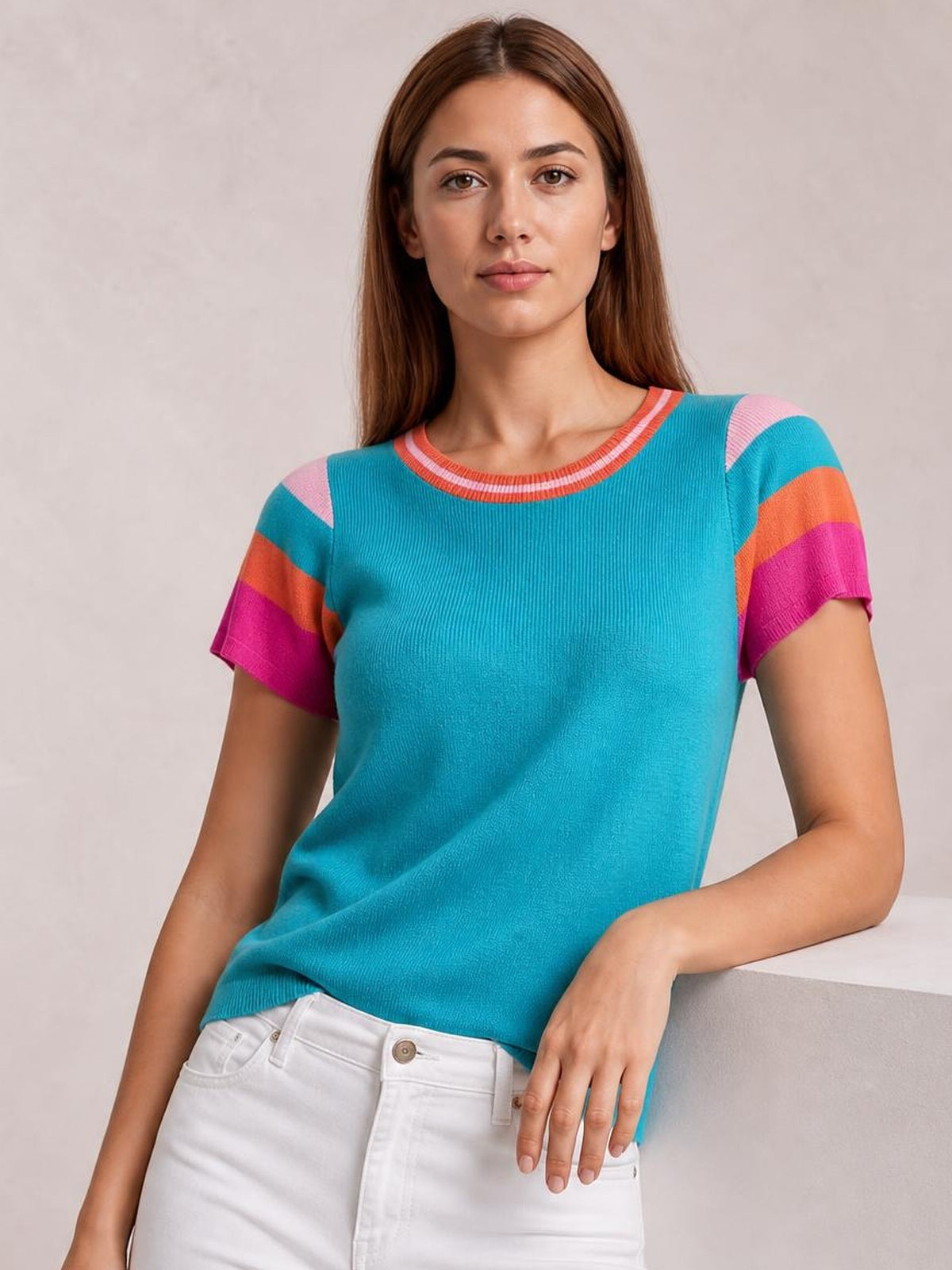 A model wears a turquoise knit top with a round neck, short multicolor striped sleeves, and a relaxed silhouette while leaning against a ledge.