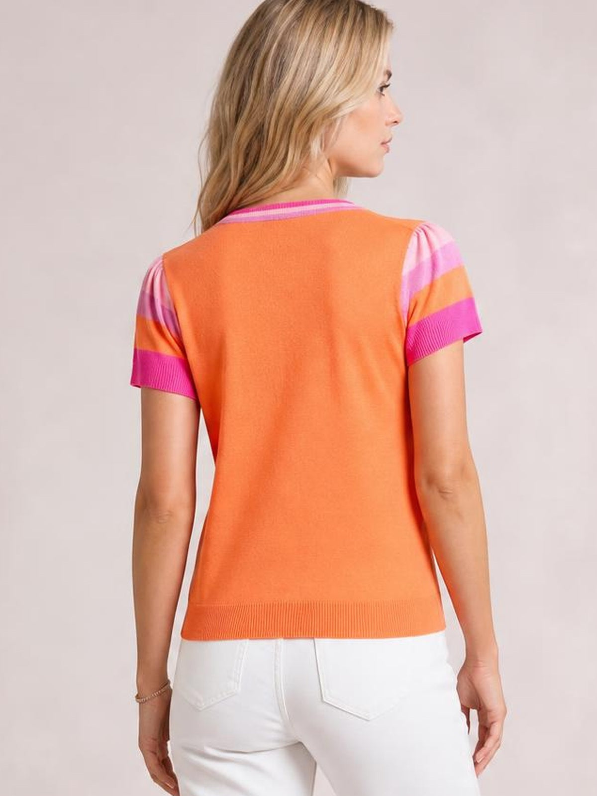 Model shown from the back wears an orange knit top with a round neck, contrast pink and lavender striped short sleeves, ribbed cuffs and hem, and a relaxed silhouette.