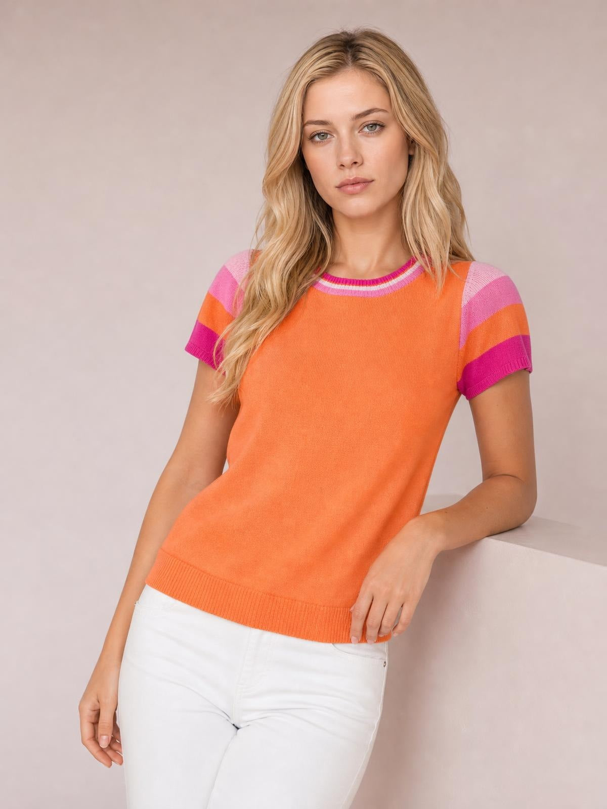 Model poses in an orange contrast knit top with a round neck, pink striped short sleeves, ribbed trim, and a relaxed silhouette.