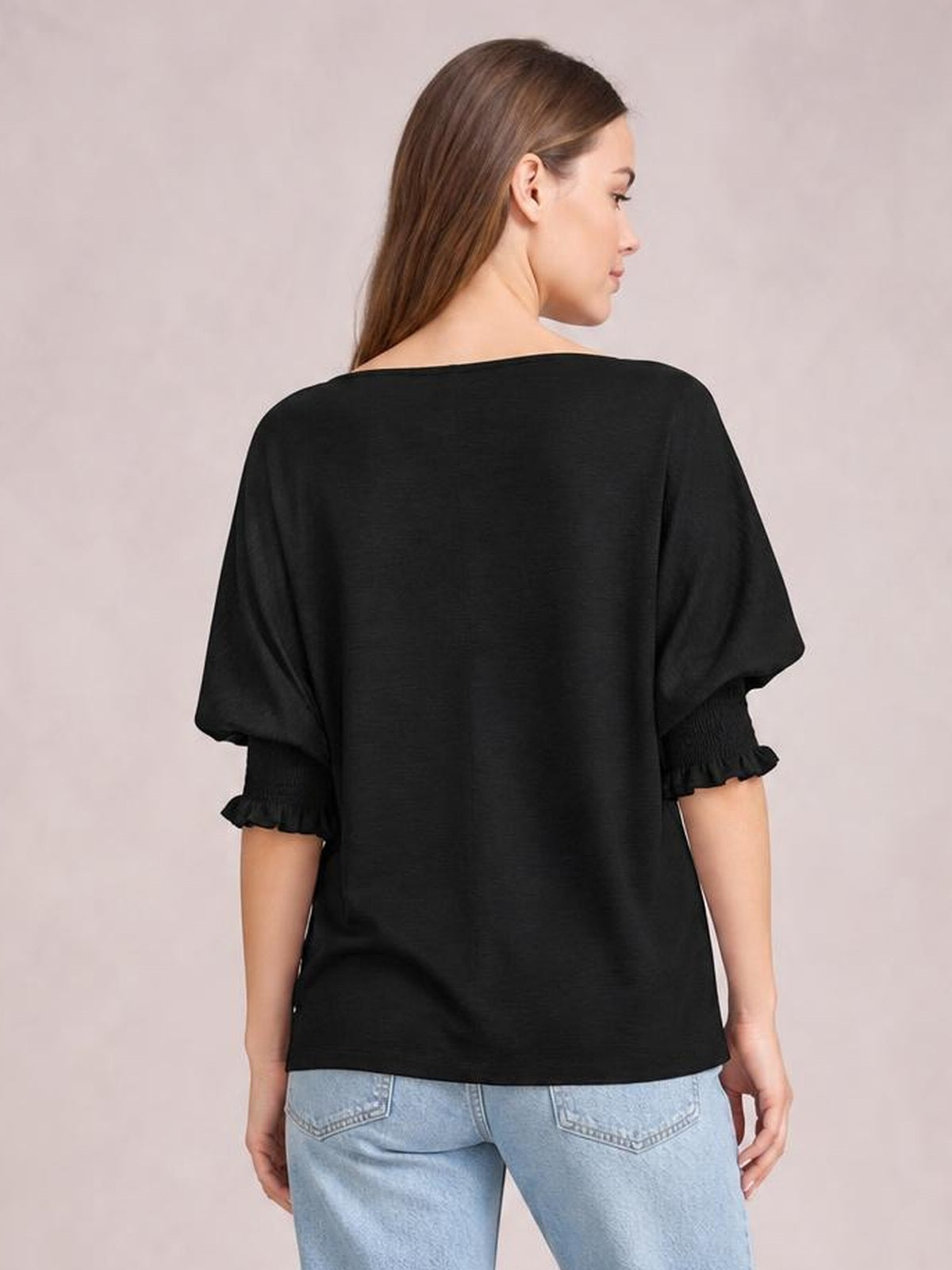 Model shown from the back in a black blouse with a loose silhouette, short lantern sleeves, and frilled cuffs, paired with light-wash jeans.