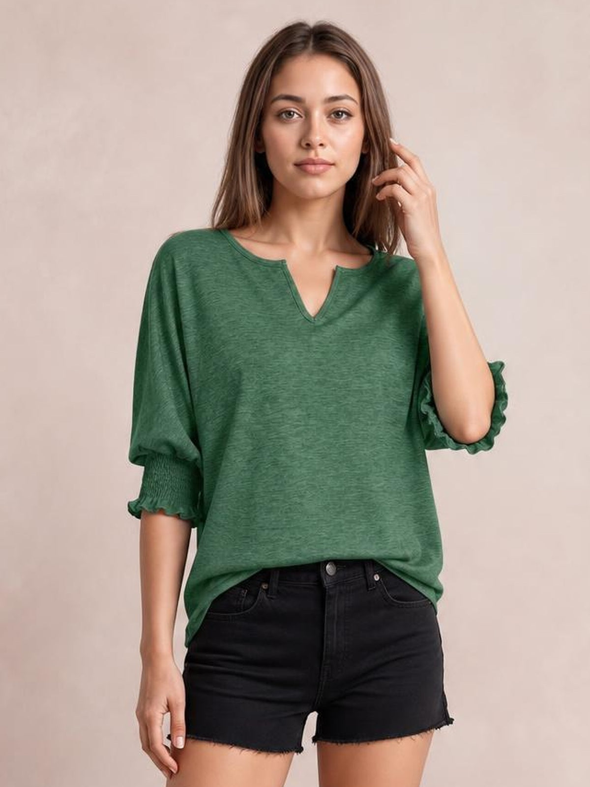 A model poses in a loose green heathered blouse with a notched neckline, lantern sleeves, and frilled cuffs, touching her hair with one hand.