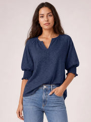 Model wears a heathered navy blouse with a notched neckline, loose silhouette, lantern sleeves, and frill-trimmed cuffs, posing with one hand in her jeans pocket.