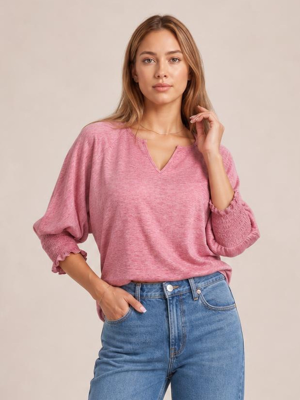 Model wears a pink heathered blouse with a notched V-neck, relaxed silhouette, lantern sleeves, and frill-trimmed cuffs, posing with one hand in her jeans pocket.