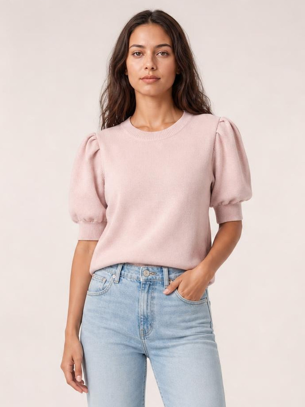 Model poses in a pale pink puff sleeve sweater with ribbed cuffs, a crew neckline, and a relaxed silhouette tucked into light-wash jeans.