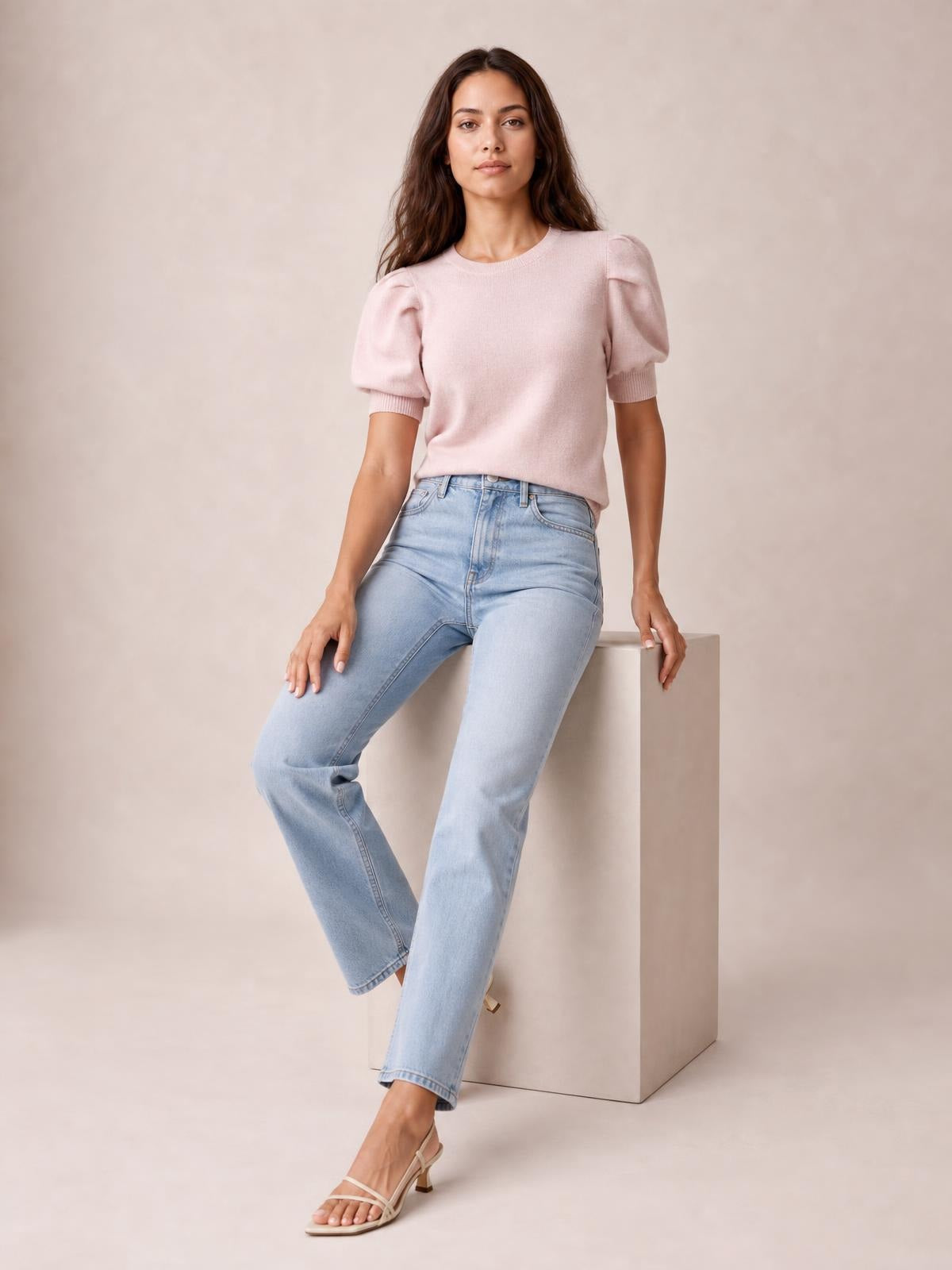 A model sits wearing a blush pink mock neck sweater with puff sleeves, ribbed cuffs, and a streamlined silhouette tucked into light-wash jeans.