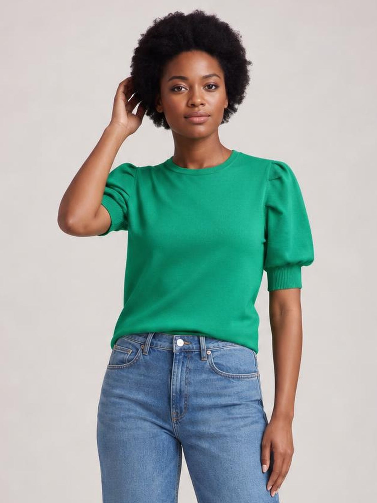 Model wearing a bright green mock neck puff sleeve sweater with a relaxed straight silhouette and ribbed sleeve cuffs, posing with one hand in her hair.