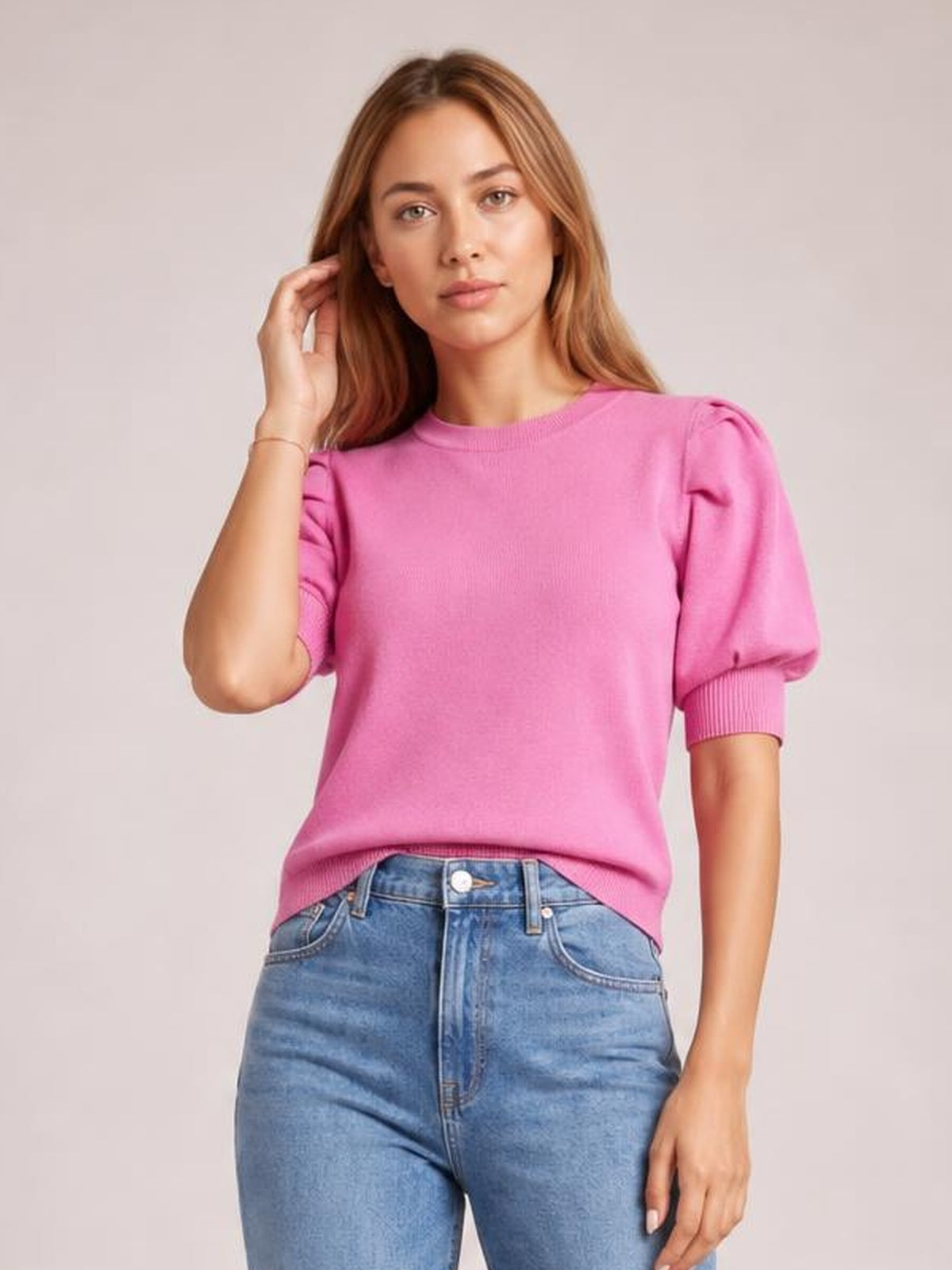 Model wearing a bright pink sweater with puffed short sleeves, ribbed cuffs and hem, a rounded neckline, and a relaxed silhouette while touching her hair.