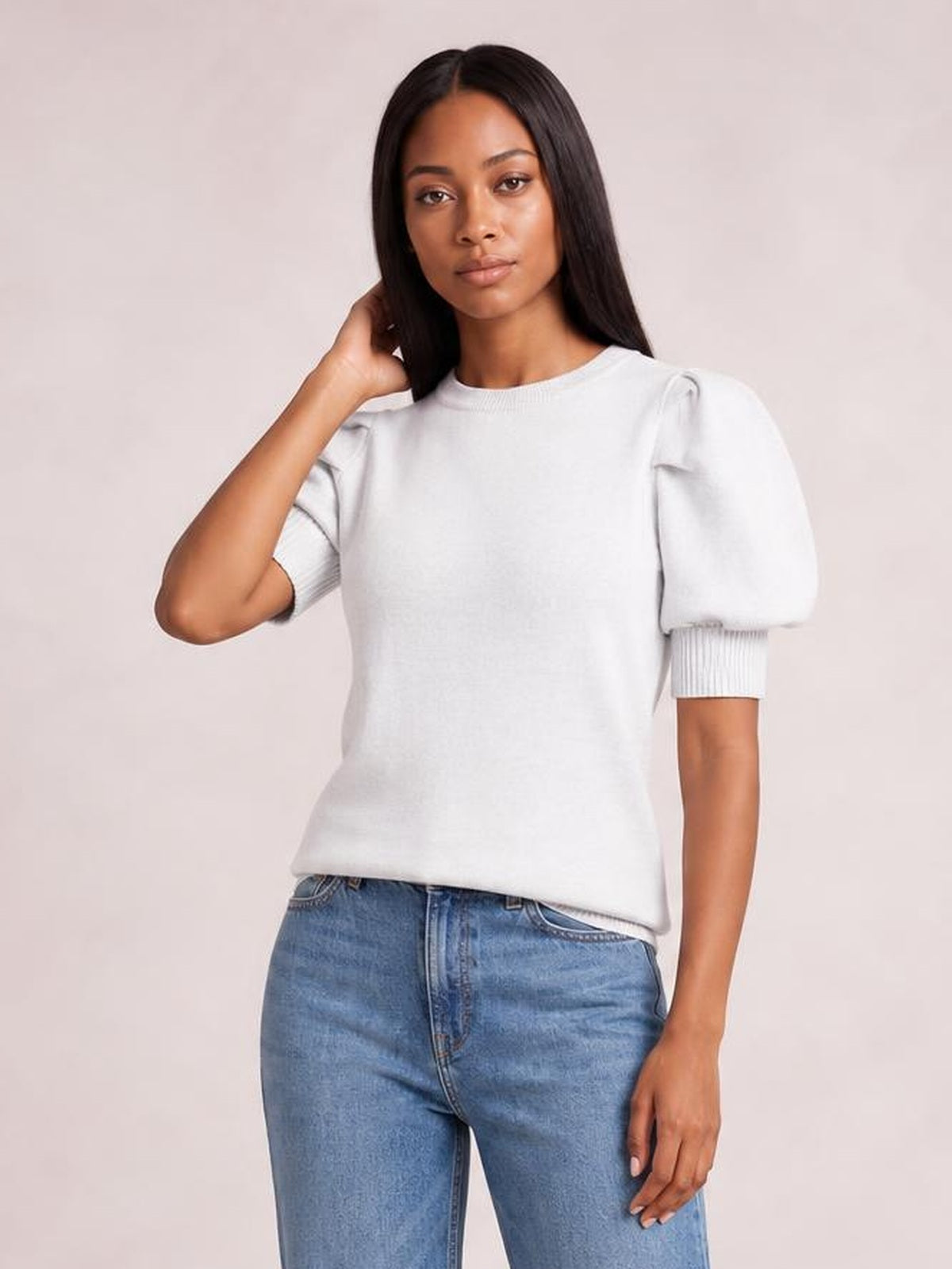 A model wears a white mock neck puff sleeve sweater with a relaxed silhouette, gathered shoulders, ribbed cuffs, and a straight hem, posing with one hand in her hair.