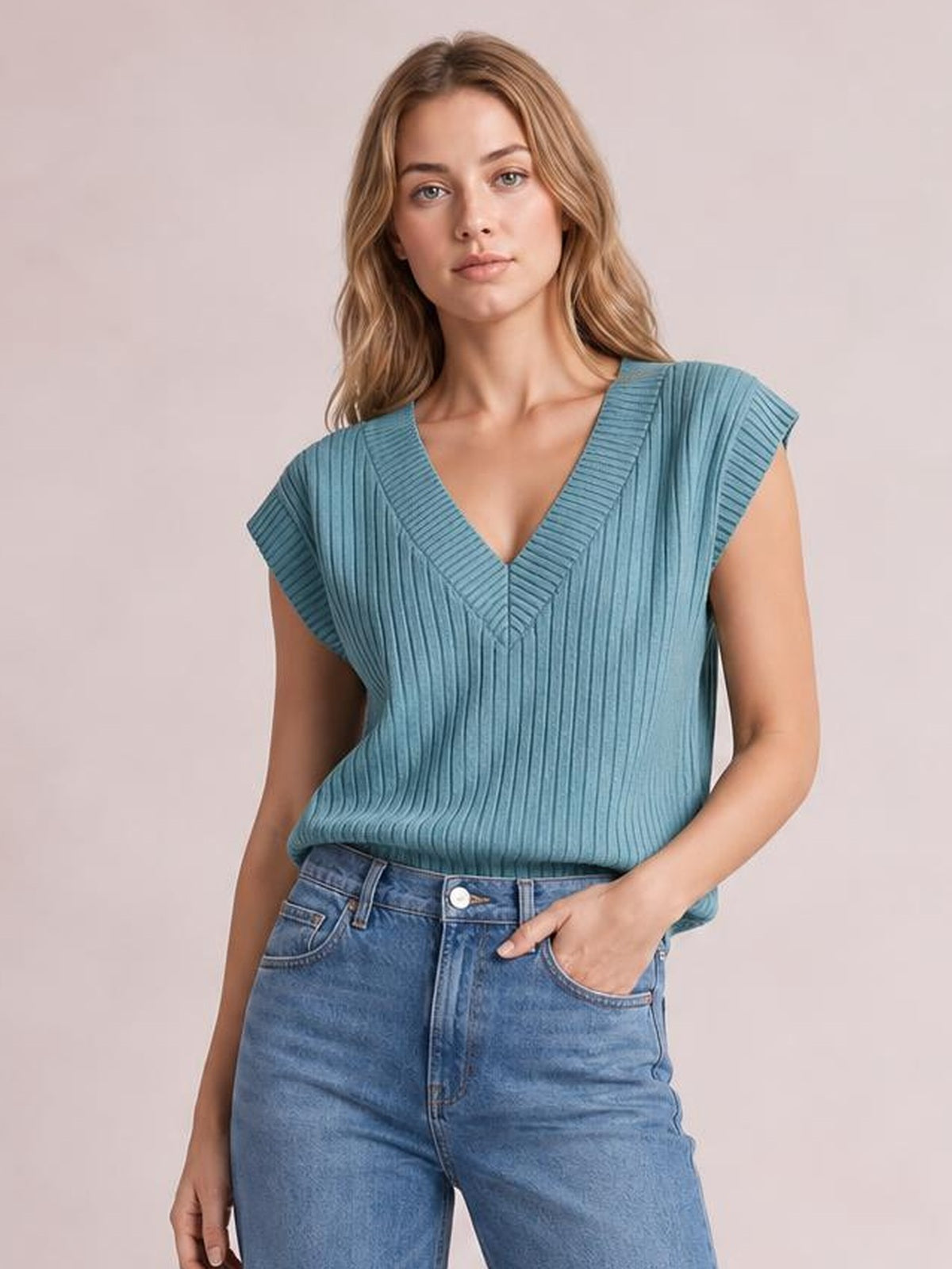 Model wearing a blue ribbed V-neck sweater vest with a relaxed sleeveless silhouette, wide shoulder panels, and a defined neckline, paired with jeans.