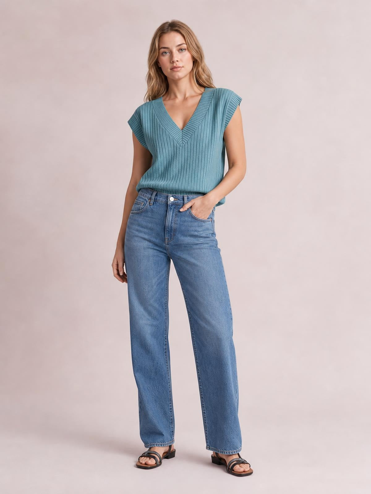 A model wears a blue ribbed V-neck sweater vest with sleeveless cap shoulders, ribbed trim, and a fitted silhouette tucked into wide-leg jeans.
