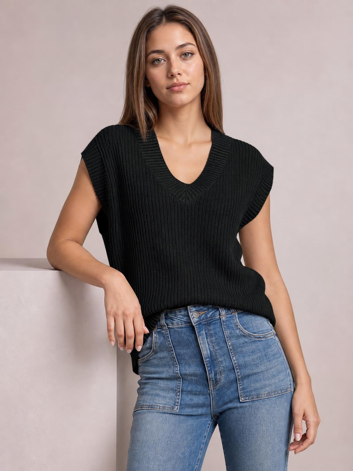 Model poses in a black ribbed V-neck sweater vest with a relaxed sleeveless silhouette, layered over jeans and resting one hand on a ledge.