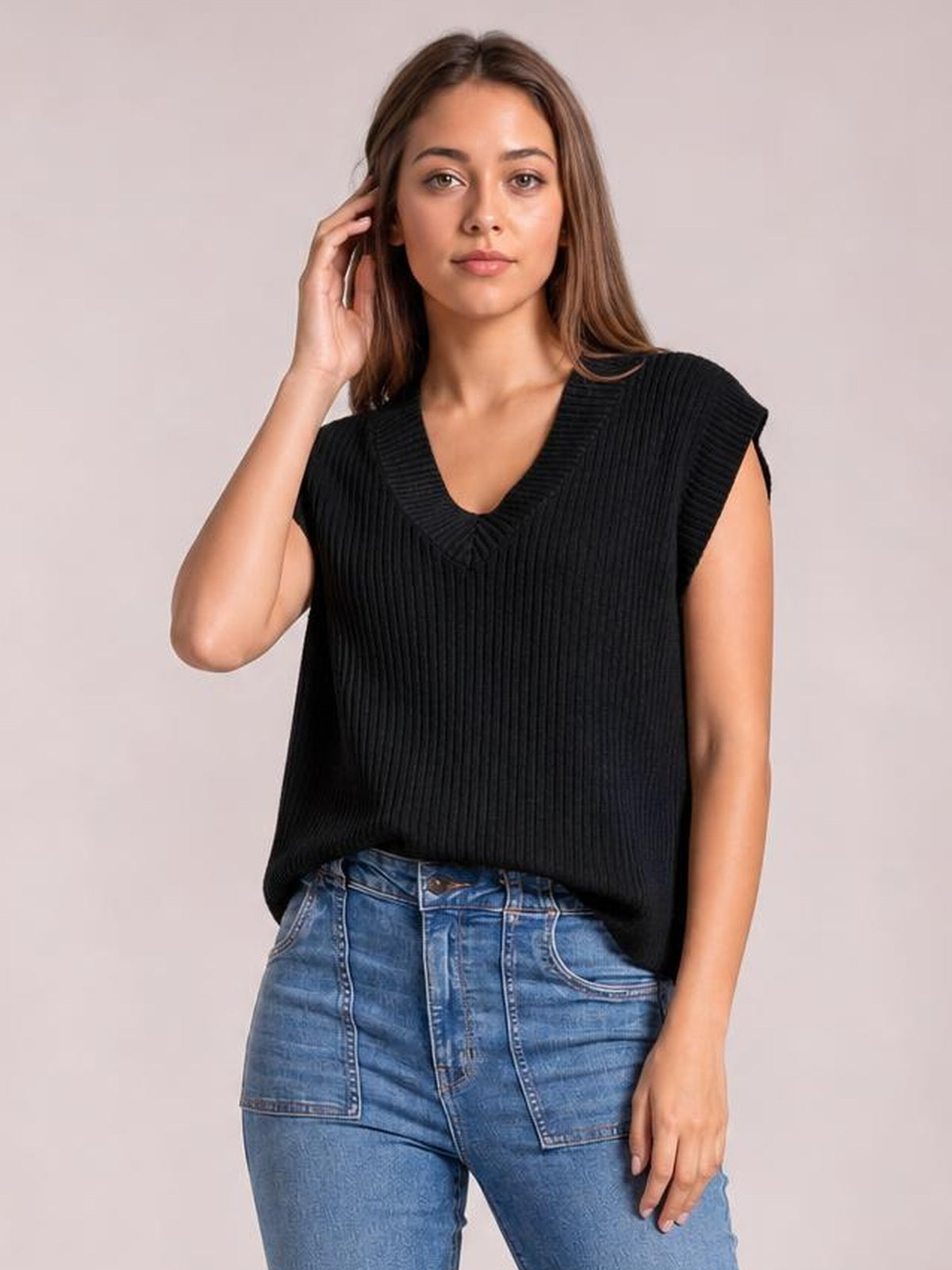 Model wearing a black ribbed V-neck sweater vest with a relaxed sleeveless silhouette, gently touching her hair while standing facing forward.