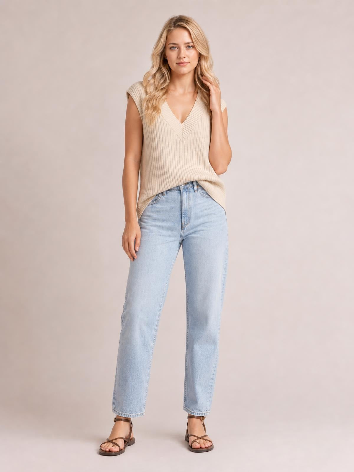 A woman stands in a cream ribbed V-neck sweater vest with a relaxed straight silhouette, sleeveless construction, and a softly extended hem over light-wash jeans.