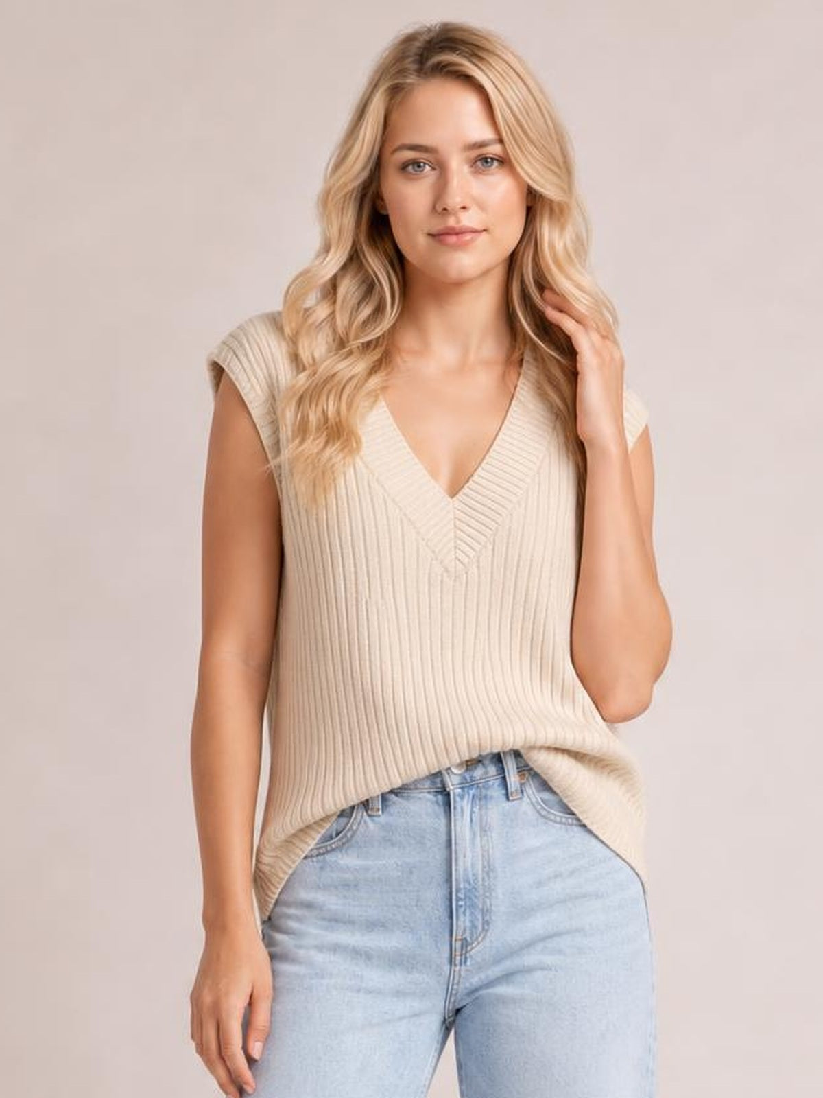 Model wearing a cream ribbed V-neck sweater vest with a relaxed sleeveless silhouette, paired with light-wash jeans while gently touching her hair.