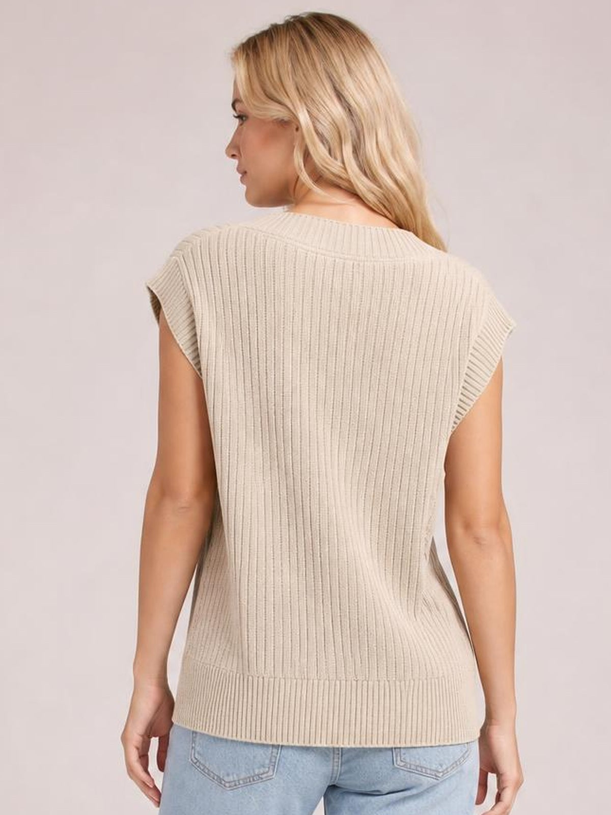 Model shown from behind wearing a beige ribbed sweater vest with sleeveless construction, a fitted straight silhouette, and a wide hem over light blue jeans.