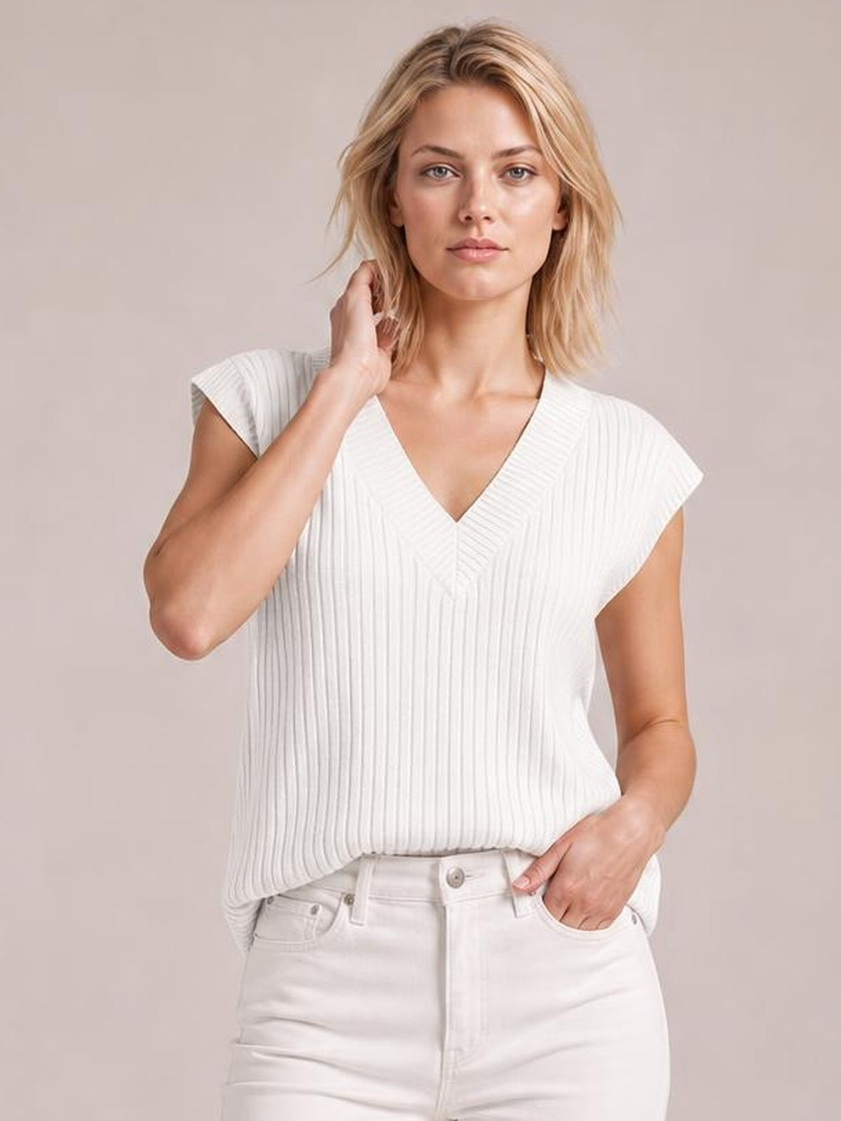 A model wears a white ribbed V-neck sweater with short sleeves and a relaxed silhouette, posing with one hand in her pocket and the other near her hair.