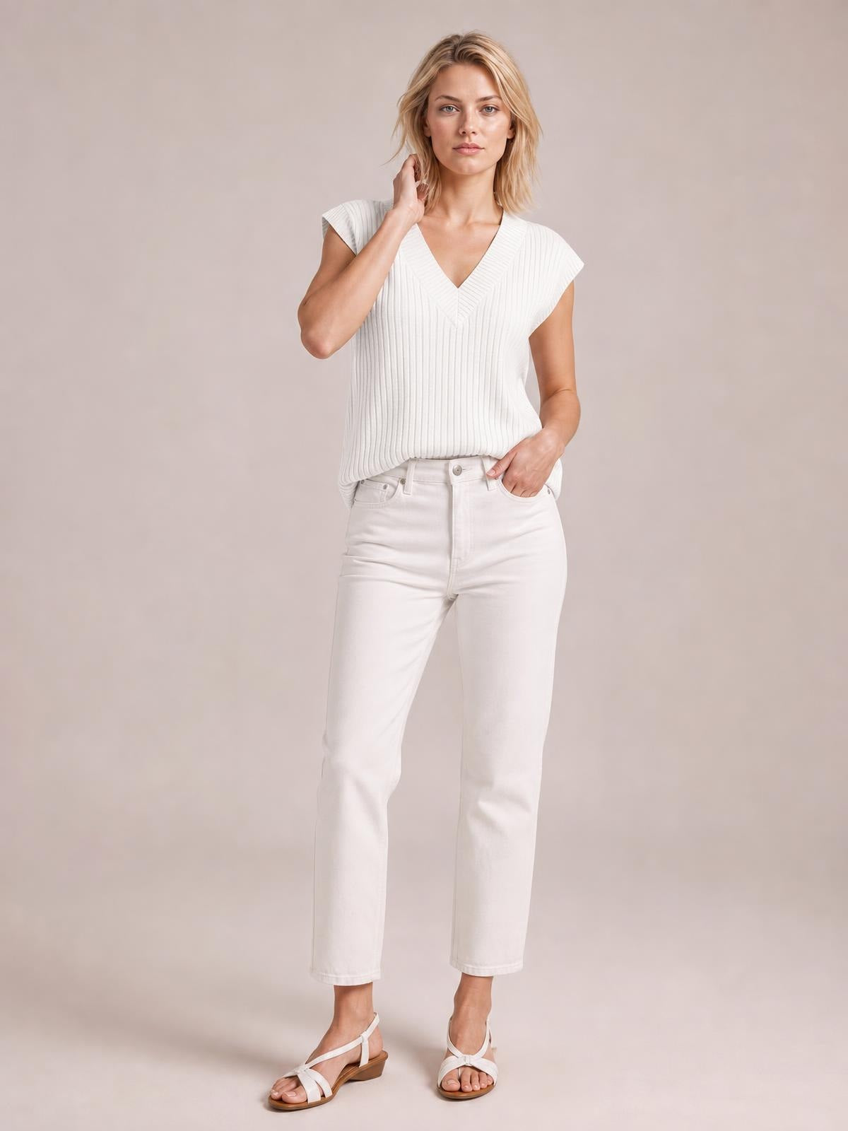 A model wears a white ribbed V-neck sweater vest with cap sleeves and a relaxed straight silhouette, standing with one hand at her neck.