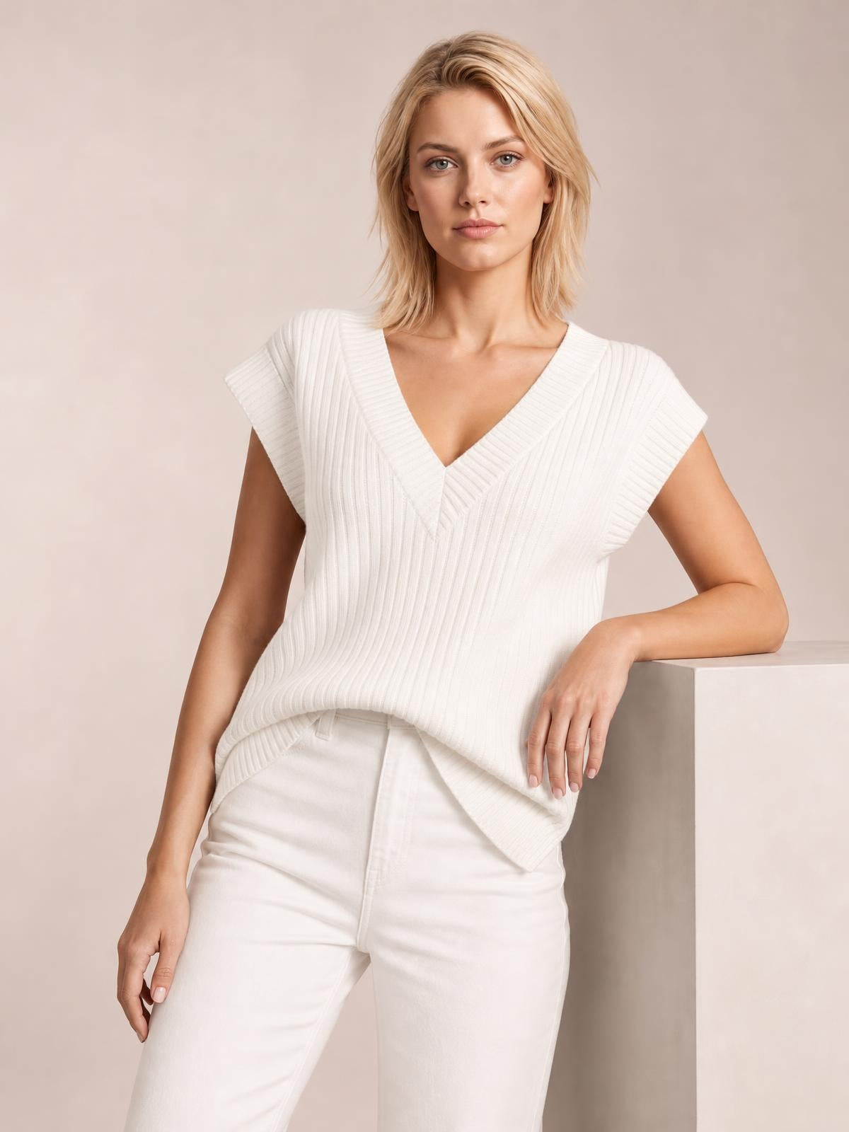 A model wears a white ribbed V-neck sweater vest with cap sleeves and a relaxed silhouette, leaning one hand on a pedestal.