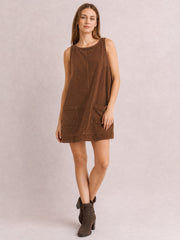 Model wears a brown round-neck mini dress with wide straps, a relaxed A-line silhouette, visible center seam, and two front patch pockets, posing with crossed legs.