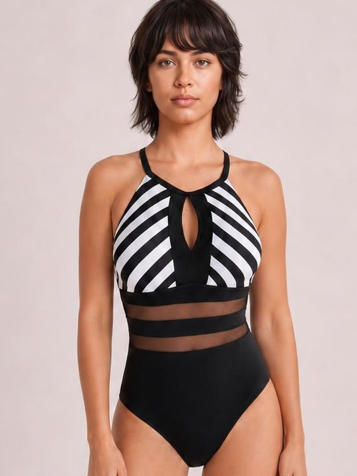 Front-facing model stands in a black and white striped one-piece swimsuit with a halter neckline, center cutout, mesh waist panels, and high-rise silhouette.