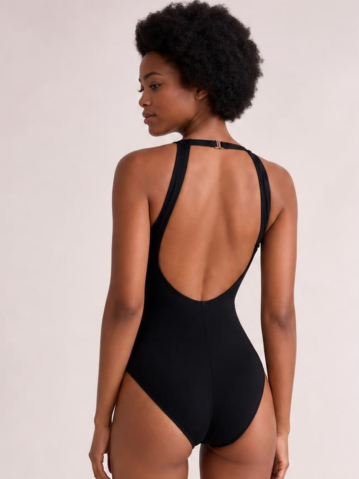 Model stands facing away in a black backless one-piece swimsuit with a deep scoop back, slim shoulder straps, high-cut leg openings, and a smooth fitted silhouette.